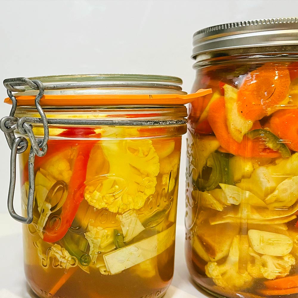 Two jars with pickled vegetables with the "In A Pickle" Mediterranean spice blend. The white cauliflower, garlic are yellowish from the healthy turmeric, along with pickled carrots, red pepper, and Jalapeños.